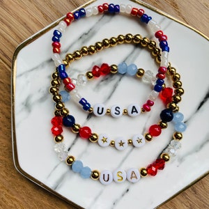May include: Three beaded bracelets with red, white and blue beads. The bracelets spell out "USA" with gold letters on white beads. The bracelets are on a white marble plate with a gold rim.