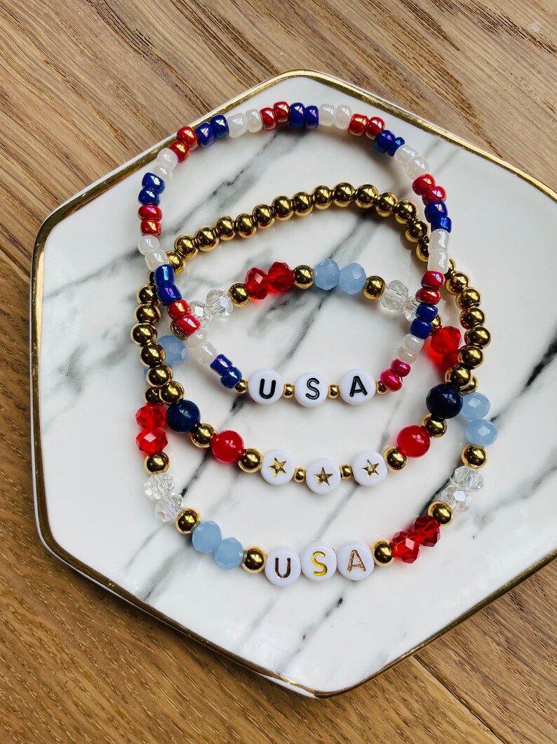 May include: Three beaded bracelets with red, white and blue beads. The bracelets spell out "USA" with gold letters on white beads. The bracelets are on a white marble plate with a gold rim.