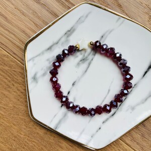 May include: A red crystal beaded bracelet with a white star charm. The bracelet is on a white marble plate with a gold rim.