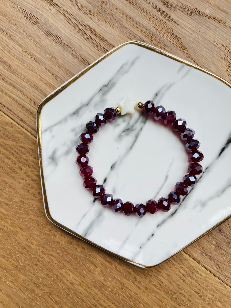 May include: A red crystal beaded bracelet with a white star charm. The bracelet is on a white marble plate with a gold rim.