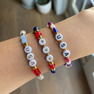 May include: Three beaded bracelets with red, white and blue beads. Each bracelet spells out "USA" with gold lettering and features a gold star charm.