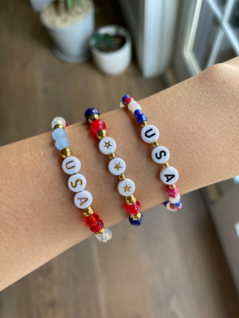 May include: Three beaded bracelets with red, white and blue beads. Each bracelet spells out "USA" with gold lettering and features a gold star charm.