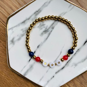May include: Gold beaded bracelet with red, white and blue beads and gold stars on the white beads.