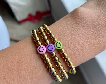 May include: Three gold beaded bracelets with smiley face beads in pink, purple, and green. The bracelets are stacked on a person's wrist, showcasing the gold beads and colorful smiley faces.