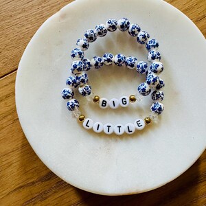 Puede incluir: Dos pulseras el&aacute;sticas con cuentas redondas de patr&oacute;n floral azul y blanco. Una pulsera incluye cuentas de letras blancas que forman la palabra BIG, mientras que la otra tiene cuentas de letras blancas que forman LITTLE, con peque&ntilde;as cuentas doradas.