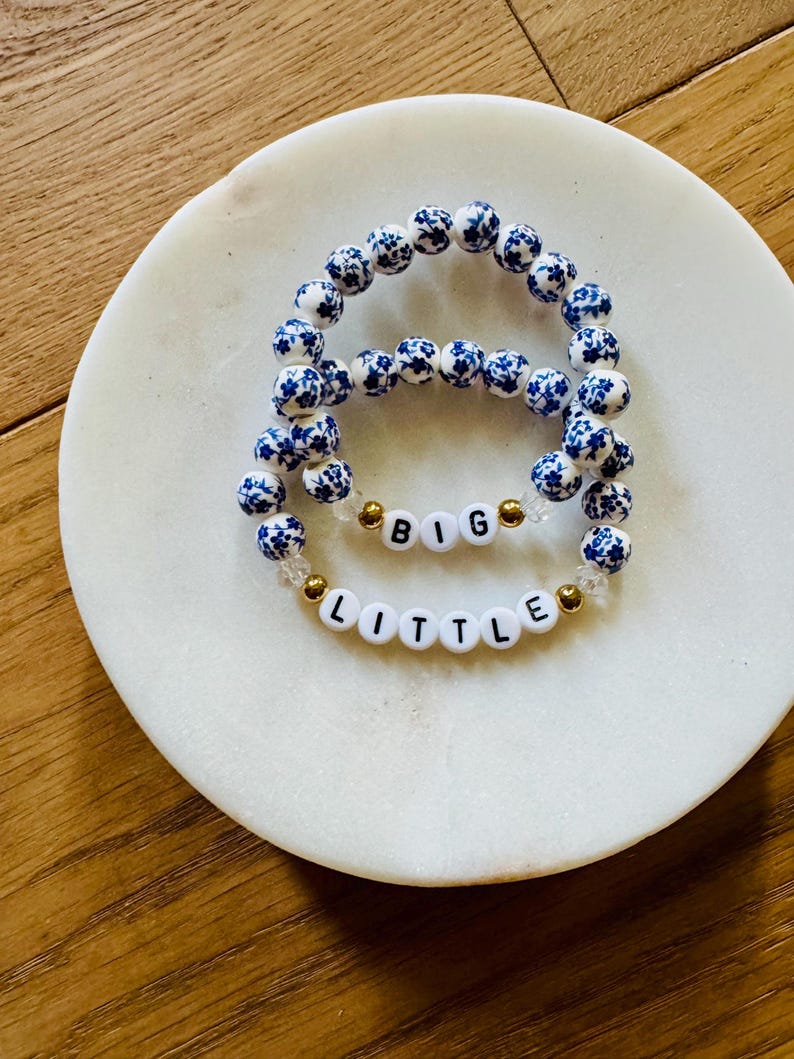 Puede incluir: Dos pulseras el&aacute;sticas con cuentas redondas de patr&oacute;n floral azul y blanco. Una pulsera incluye cuentas de letras blancas que forman la palabra BIG, mientras que la otra tiene cuentas de letras blancas que forman LITTLE, con peque&ntilde;as cuentas doradas.