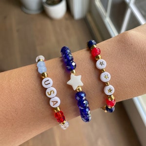 May include: Three beaded bracelets in red, white and blue. One bracelet has blue beads and a white star charm. Another bracelet has red and white beads and a white star charm. The third bracelet has white beads with the letters "USA" in gold.