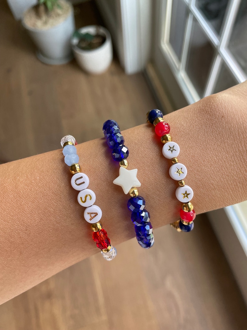 May include: Three beaded bracelets in red, white and blue. One bracelet has blue beads and a white star charm. Another bracelet has red and white beads and a white star charm. The third bracelet has white beads with the letters "USA" in gold.