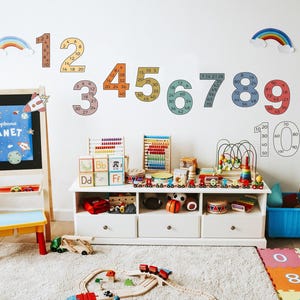 Op de afbeelding: Een kleurrijke speelkamer met een witte muur die is versierd met regenboog- en cijferdecals. De cijfers zijn gevuld met kleinere cijfers, tellend tot 20. Een giraffen-hoogtekaart hangt aan de muur. Er is een houten ezel met een schoolbord, een houten tafel met speelgoed en een blauwe bak. De vloer is bedekt met kleurrijke schuimpuzzelmatten.