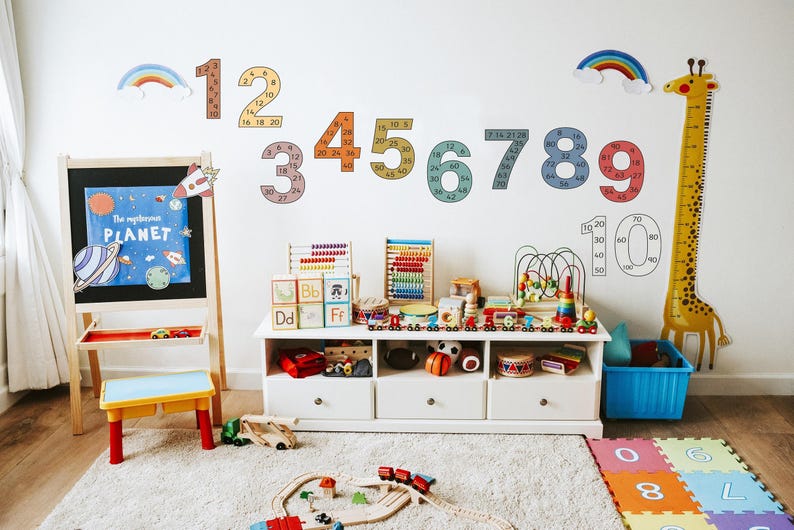 Op de afbeelding: Een kleurrijke speelkamer met een witte muur die is versierd met regenboog- en cijferdecals. De cijfers zijn gevuld met kleinere cijfers, tellend tot 20. Een giraffen-hoogtekaart hangt aan de muur. Er is een houten ezel met een schoolbord, een houten tafel met speelgoed en een blauwe bak. De vloer is bedekt met kleurrijke schuimpuzzelmatten.