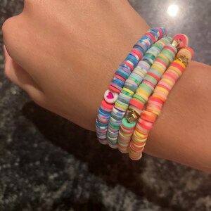 May include: A collection of colorful beaded bracelets. The bracelets feature a variety of colors, including pink, blue, yellow, and green. Some bracelets have heart and star charms. The beads are flat and round.
