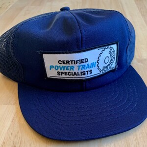 May include: A navy blue trucker hat with a mesh back. The hat has a white patch with the text "CERTIFIED POWER TRAIN SPECIALISTS" and a blue gear graphic.