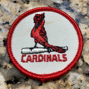 May include: A red, white, and gray embroidered patch with a red cardinal perched on a baseball bat. The text "CARDINALS" is embroidered in red below the cardinal.
