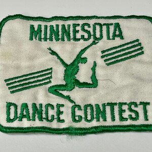 Pode incluir: Um remendo de tecido branco com texto bordado em verde e uma silhueta verde de um dançarino. O texto diz "Minnesota Dance Contest".