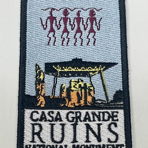 Könnte beinhalten: Ein gestickter Aufnäher in Schwarzweiß mit dem Text "Casa Grande Ruins National Monument". Der Aufnäher zeigt ein stilisiertes Bild eines Gebäudes mit einem rot-weiß-blauen Design und vier Strichmännchen in Rot.