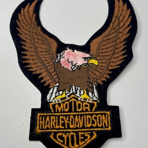 Może przedstawiać: Naszywka haftowana przedstawiająca brązowego orła z rozpostartymi skrzydłami, usadowionego na czarno-złotym logo Harley Davidson z napisem "MOTOR HARLEY DAVIDSON CYCLES".
