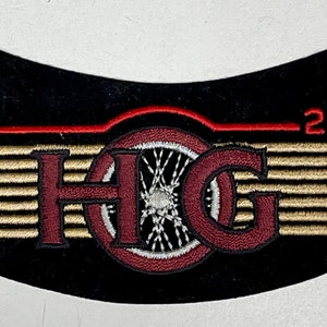 Peut inclure: Un écusson noir incurvé avec des ailes brodées en or et rouge, un cercle rouge avec une roue blanche à l'intérieur et les lettres "HOG" en rouge avec un contour doré. Le texte "2001" est brodé en rouge au-dessus des ailes.