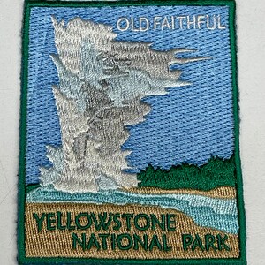 Op de afbeelding: Geborduurde patch met een geiser die uitbarst tegen een blauwe lucht, met de tekst "OLD FAITHFUL" bovenaan. De onderkant van de patch luidt "YELLOWSTONE NATIONAL PARK" in groen en goud. De patch is groen omrand.