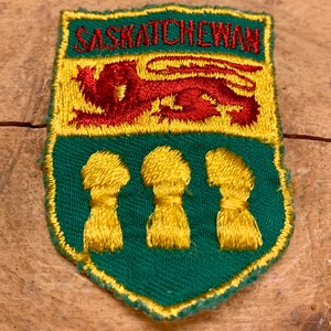 May include: A green and yellow embroidered patch with the word "Saskatchewan" in white lettering. The patch features a red lion and three yellow wheat sheaves.