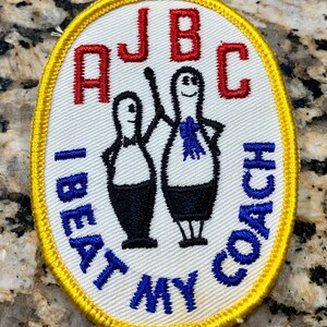 Puede incluir: Un parche ovalado blanco con un borde amarillo. El parche tiene texto azul que dice "I Beat My Coach" y texto rojo que dice "AJBC". Dos bolos negros y blancos están en el centro del parche.