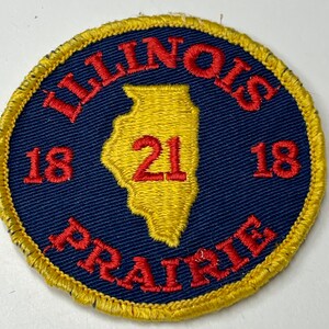 Könnte beinhalten: Ein gestickter Aufnäher in Blau und Gelb mit dem Text "ILLINOIS PRAIRIE" und der Zahl "21" innerhalb einer Umrandung des Bundesstaates Illinois. Die Zahlen "18" befinden sich auf beiden Seiten der Bundesstaatenumrandung.