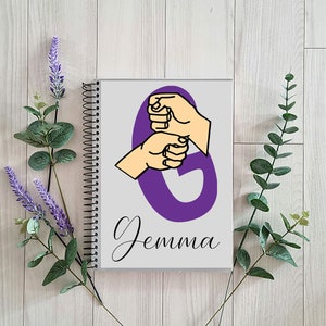 May include: Purple spiral notebook with a purple letter G and two hands forming the letter G in sign language. The notebook has the name "Gemma" written on it.