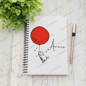 May include: A white spiral bound notebook with a black and white illustration of a bear holding a red balloon. The name "Annie" is written in cursive on the notebook.