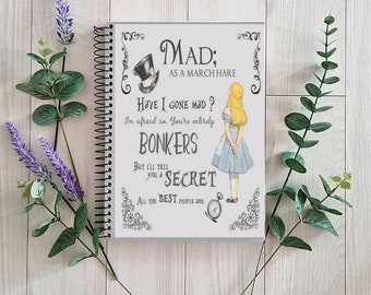 May include: A spiral bound notebook with a white cover featuring a black and white illustration of Alice from Alice in Wonderland. The text on the cover reads "MAD; AS A MARCH HARE HAVE I GONE MAD? I'm afraid so. You're entirely BONKERS BUT I'LL TELL you a SECRET ALL THE BEST PEOPLE ARE"