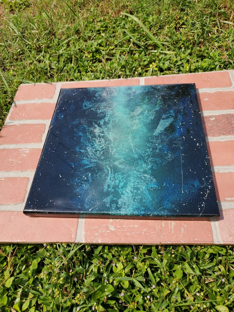 May include: A square canvas painting with a blue and black abstract design. The painting has a white splatter effect and a light blue gradient.