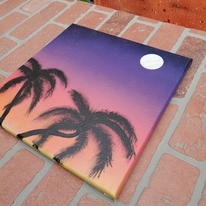 May include: A hand-painted canvas with a sunset scene featuring two palm trees silhouetted against a gradient background of orange, yellow, pink, and purple. A white moon is visible in the upper right corner of the canvas.