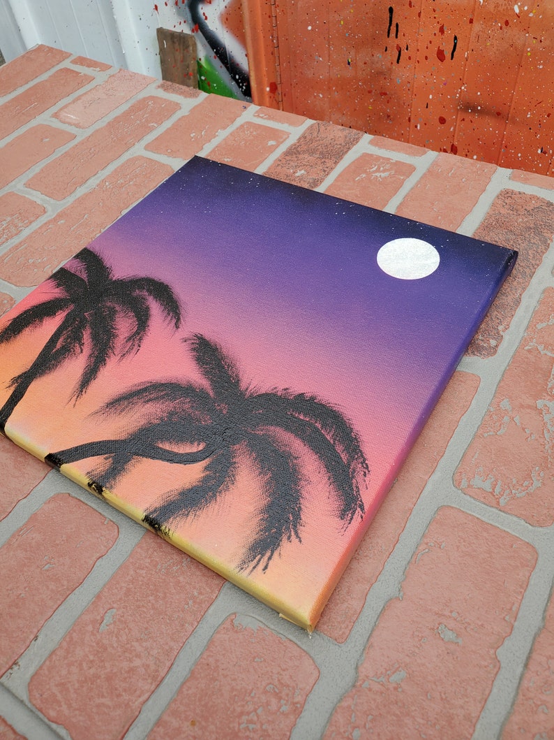 May include: A hand-painted canvas with a sunset scene featuring two palm trees silhouetted against a gradient background of orange, yellow, pink, and purple. A white moon is visible in the upper right corner of the canvas.