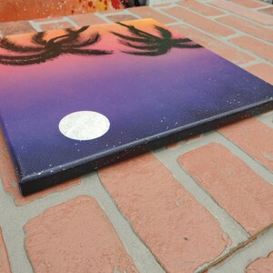 May include: A painting on canvas depicting a sunset with palm trees silhouetted against a purple and orange sky. A white moon is visible in the sky.