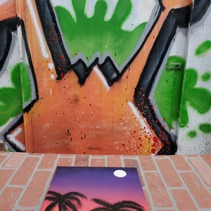 May include: A hand-painted canvas with a sunset scene featuring two palm trees and a full moon. The background is a gradient of purple, pink, and orange.