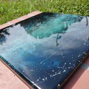 May include: A square canvas painting with a deep blue and green abstract design. The painting has a glossy finish and white speckles throughout.