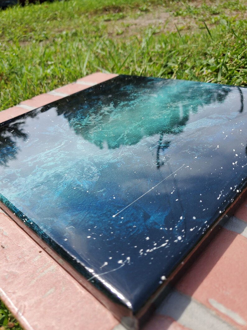 May include: A square canvas painting with a deep blue and green abstract design. The painting has a glossy finish and white speckles throughout.