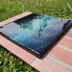 May include: A square canvas painting with a black background and a swirling green and blue design. The painting has a glossy finish and white glitter embedded in the paint.