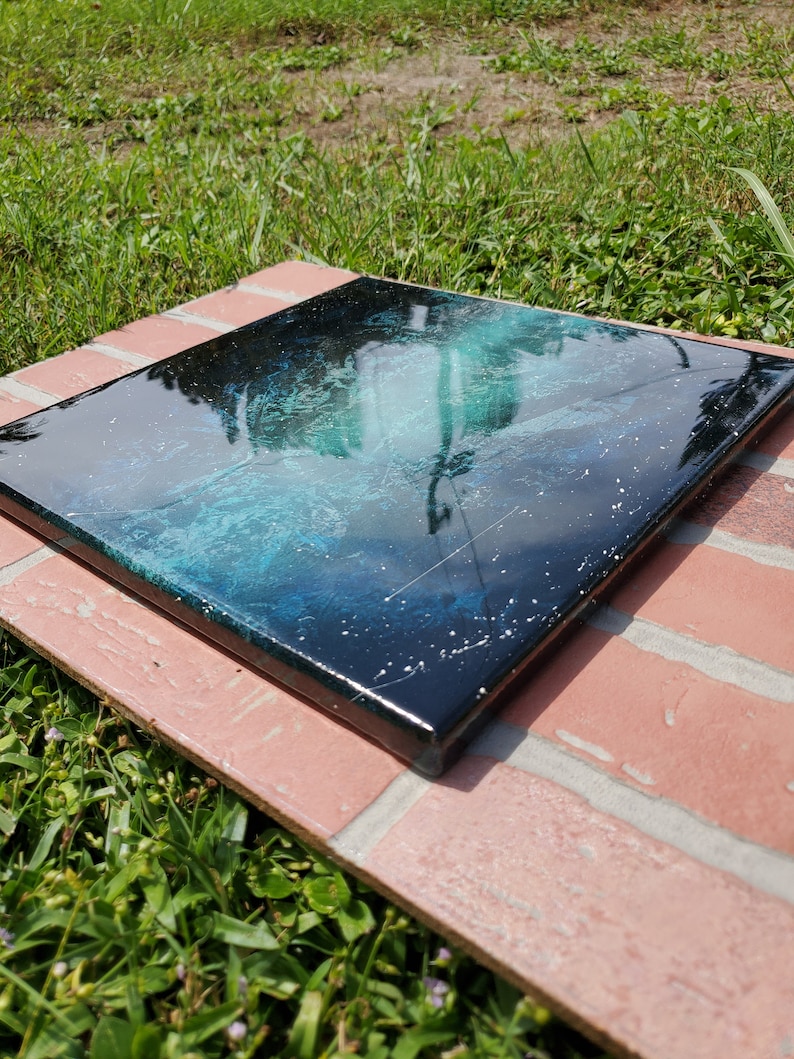 May include: A square canvas painting with a black background and a swirling green and blue design. The painting has a glossy finish and white glitter embedded in the paint.