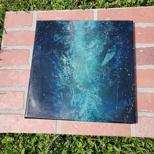 May include: A square canvas painting with a blue and black abstract design. The painting has a white splatter effect and a light blue gradient.