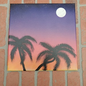 May include: A painting of a sunset with two palm trees silhouetted against the orange and pink sky. A full moon is visible in the upper right corner of the painting.