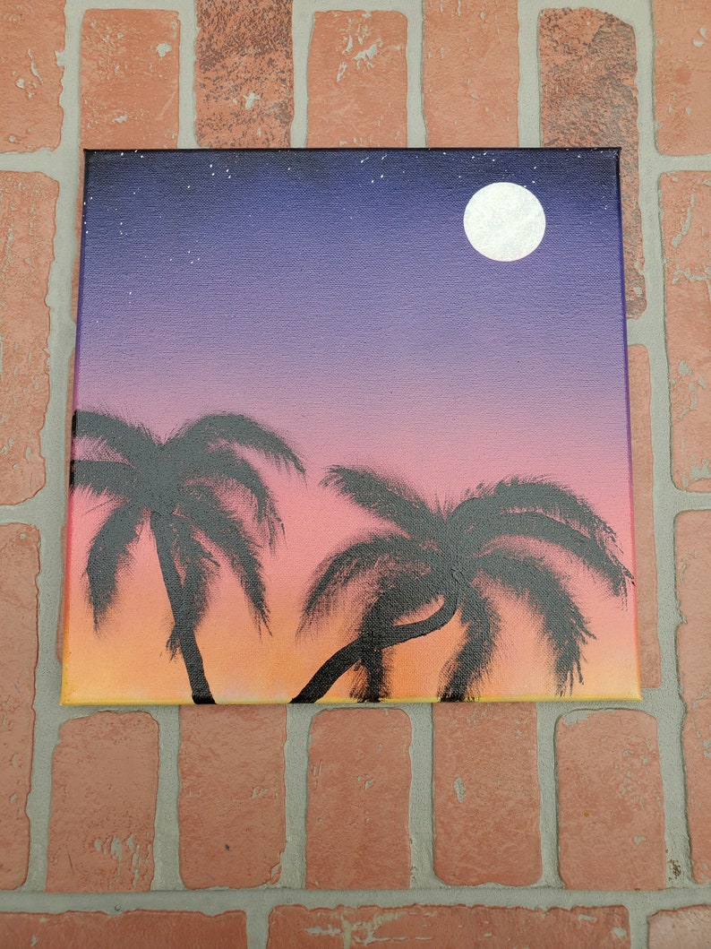 May include: A painting of a sunset with two palm trees silhouetted against the orange and pink sky. A full moon is visible in the upper right corner of the painting.