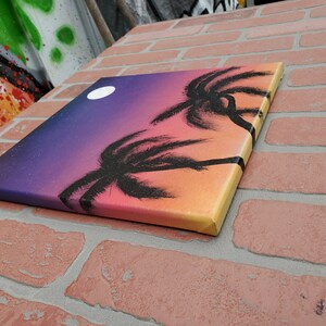 May include: A canvas painting with a sunset gradient background in shades of purple, pink, orange, and yellow. Two black palm trees are silhouetted against the sunset. A white moon is visible in the upper left corner of the painting.