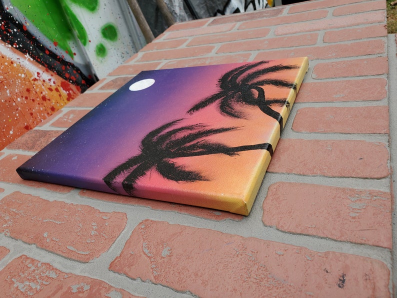 May include: A canvas painting with a sunset gradient background in shades of purple, pink, orange, and yellow. Two black palm trees are silhouetted against the sunset. A white moon is visible in the upper left corner of the painting.