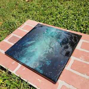 May include: A square canvas painting with a black background and a swirling blue and green nebula design. The painting has white speckles throughout, resembling stars.