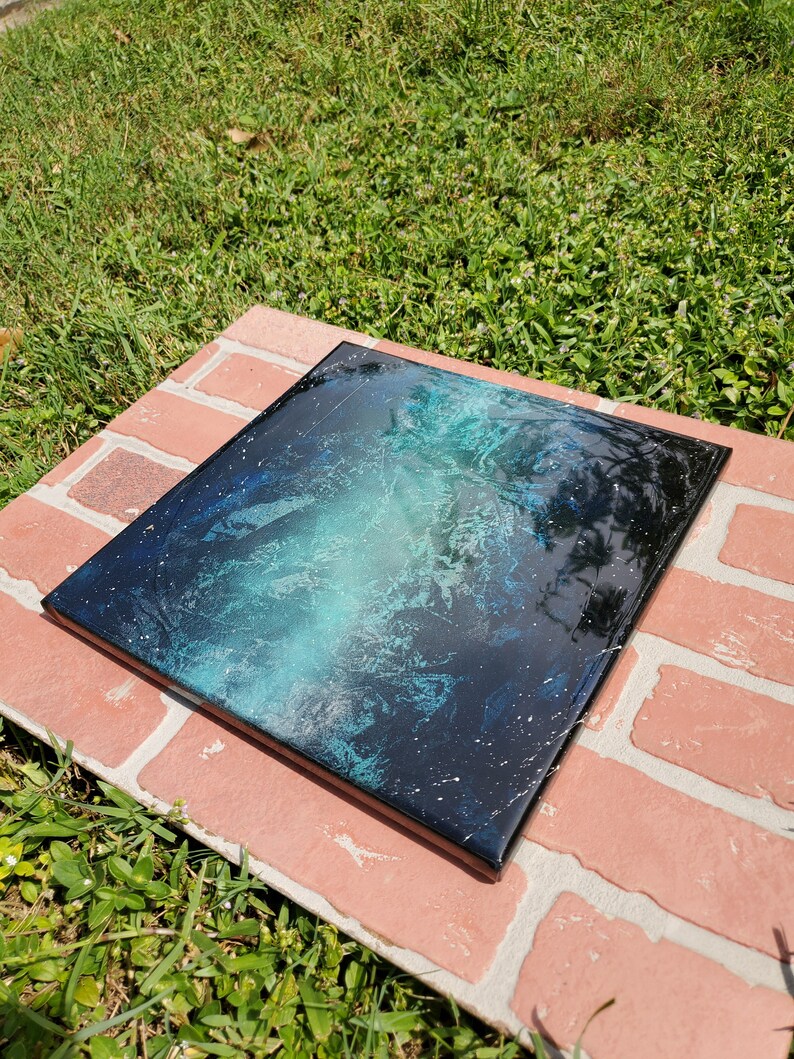 May include: A square canvas painting with a black background and a swirling blue and green nebula design. The painting has white speckles throughout, resembling stars.