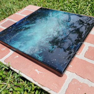 May include: A square canvas painting with a black background and a blue and white abstract design. The design resembles a galaxy with white stars and a blue nebula.