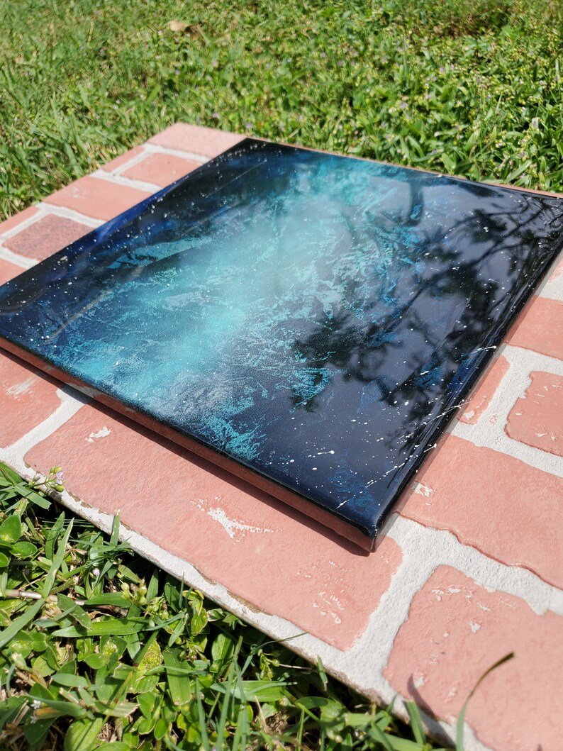 May include: A square canvas painting with a black background and a blue and white abstract design. The design resembles a galaxy with white stars and a blue nebula.