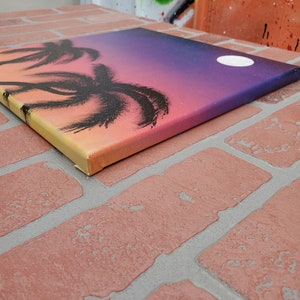 May include: A painting on canvas depicting a sunset with palm trees silhouetted against the orange, pink, and purple sky. A white moon is visible in the upper right corner of the painting.