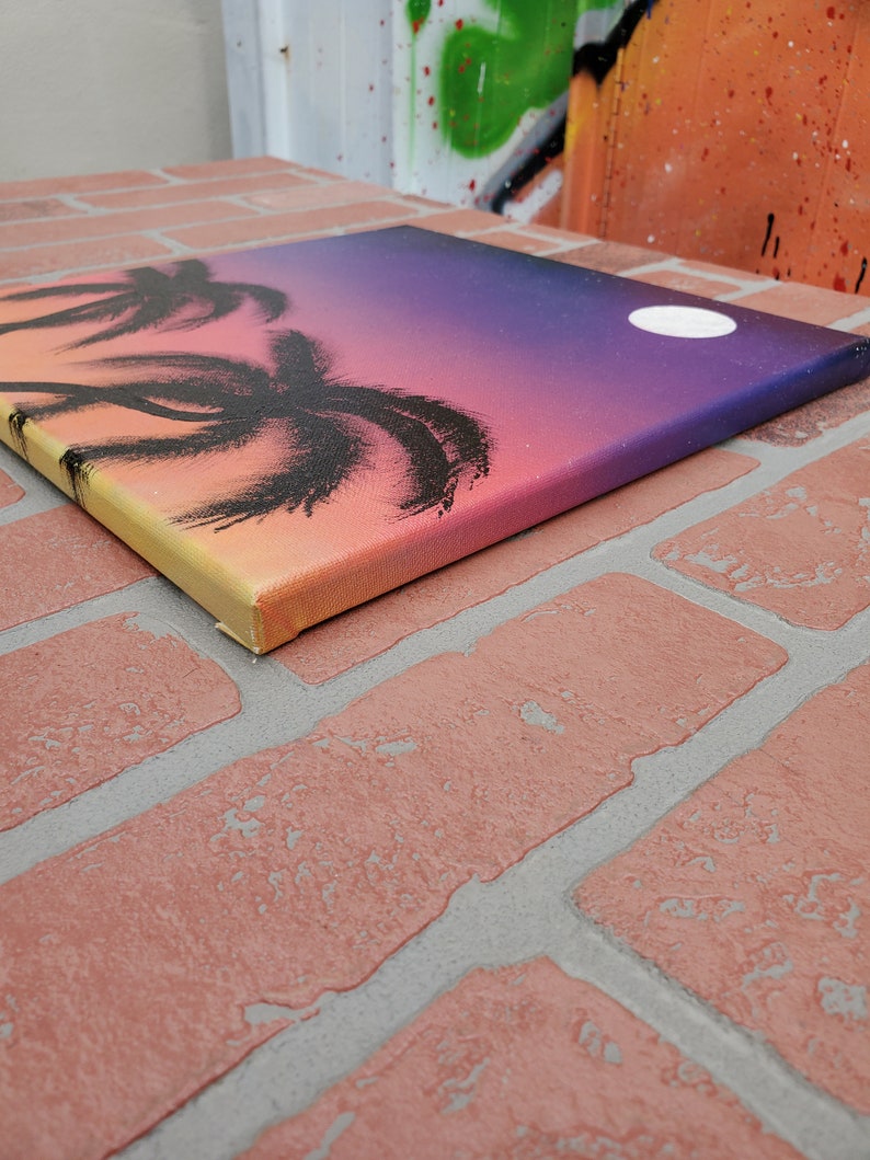 May include: A painting on canvas depicting a sunset with palm trees silhouetted against the orange, pink, and purple sky. A white moon is visible in the upper right corner of the painting.