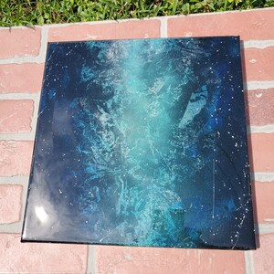 May include: A square canvas painting with a blue and green abstract design. The background is black with white speckles.