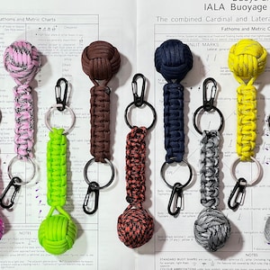 Peut inclure: Sept porte-clés en paracorde de couleurs différentes avec un nœud à l'extrémité. Les porte-clés sont de couleurs différentes, notamment rose, vert, marron, bleu, jaune et gris. Les porte-clés sont suspendus à un tableau avec des symboles nautiques et du texte.